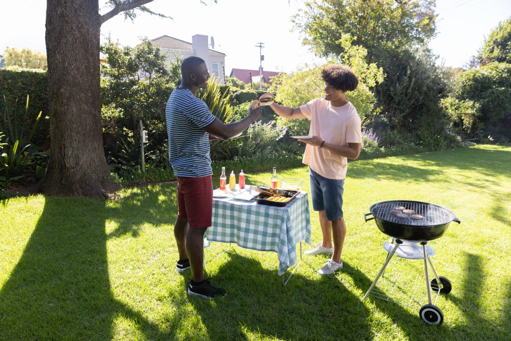 Two black men tapping their burgers against each other at a backyard picnic table on a green, well-cared-for lawn.